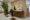 A modern salon with two black and gold styling chairs, wooden stations with round mirrors, beauty equipment, and decorative plants. The space has light-colored walls and floors, creating a clean and stylish atmosphere.