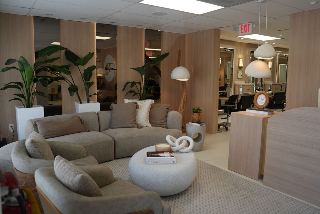 Modern waiting area with a gray sectional sofa, round ottoman, neutral decor, potted plants, and soft lighting. Large mirrors and wooden accents create an inviting, contemporary atmosphere. Office or salon workstations are visible in the background.