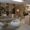 Modern waiting area with a gray sectional sofa, round ottoman, neutral decor, potted plants, and soft lighting. Large mirrors and wooden accents create an inviting, contemporary atmosphere. Office or salon workstations are visible in the background.