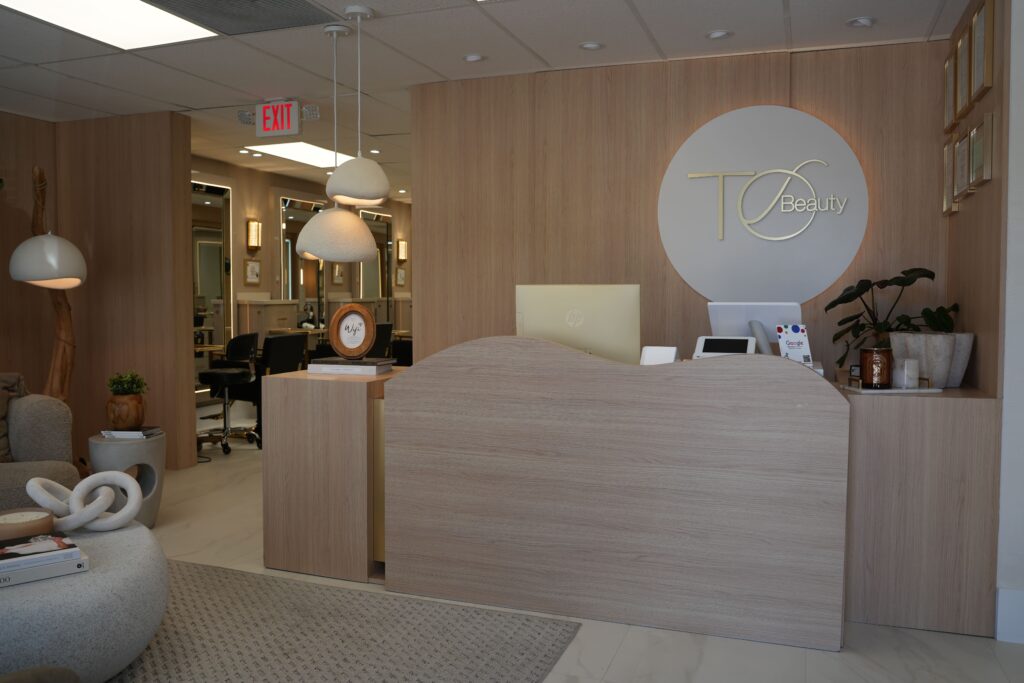 A modern beauty salon reception area with light wood decor, a curved front desk, pendant lights, plants, and a TC Beauty logo on the wall. Salon stations with mirrors are visible in the background.