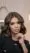 A woman with wavy, shoulder-length brown hair and highlights—fresh from a beauty salon—poses indoors in a black top and blazer. With subtle makeup, nude lipstick, manicured nails, and a forearm tattoo, she looks confidently at the camera.