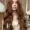 A young woman with long, wavy auburn hair stands indoors wearing a cream-colored blouse and blue jeans, looking directly at the camera—her style is perfect inspiration for your next visit to a hair salon near me. Light wood paneling and a white lamp set the scene.