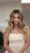 A woman with long, wavy, blonde hair styled by expert hair stylists near me wears a strapless white dress indoors, looking confidently at the camera. The softly blurred background highlights her as the main focus.