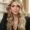 A woman with long, wavy blonde hair wearing a black outfit and gold necklace poses indoors at a beauty salon, touching her hair. Shelves with products are visible in the blurred background.