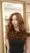A woman with long, wavy auburn hair and blue eyes stands indoors, wearing a black off-shoulder top. She looks confidently at the camera—her style and poise are reminiscent of a visit to a top beauty salon or hair stylists near me. Shelves and a mirror glow softly behind her.