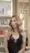 A woman with long, wavy blonde hair wearing a black top stands indoors, looking slightly to the side. Shelves with beauty products are blurred in the background of a vibrant hair salon near me.