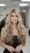 A woman with long, wavy blonde hair wearing a black strapless top stands indoors in a well-lit hair salon, looking directly at the camera with a neutral expression.